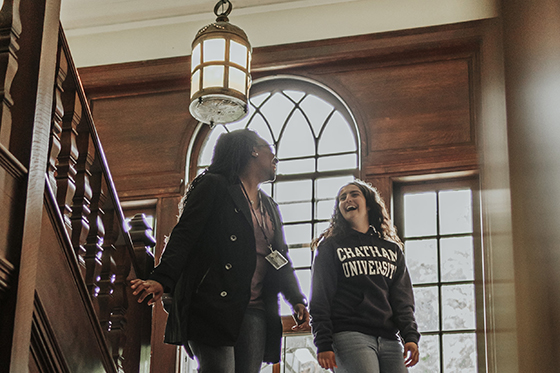 Photo of two female Chatham University students walk down wooden stairwell talking and laughing together.